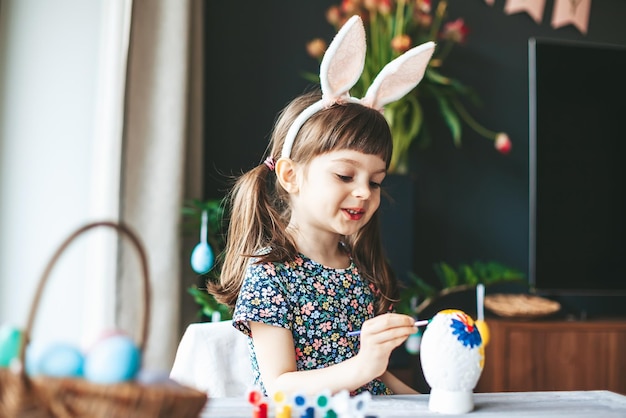 Adorable little girl with bunny ears painting gypsum Easter egg with paintbrush