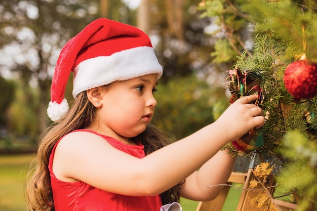 Adorable little girl with Christmas hat puts a ball on the Christmas tree