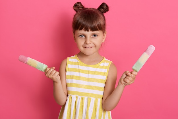 Adorable little girl with two knots holding ice cream isolated over rose background, wearing summer dress, looking at camera, having curious expression.