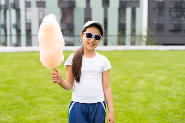 Adorable and pretty curly little girl eating sweet cotton candy. Happy child, kid eating candy-floss with emotions in the park at summer or spring.