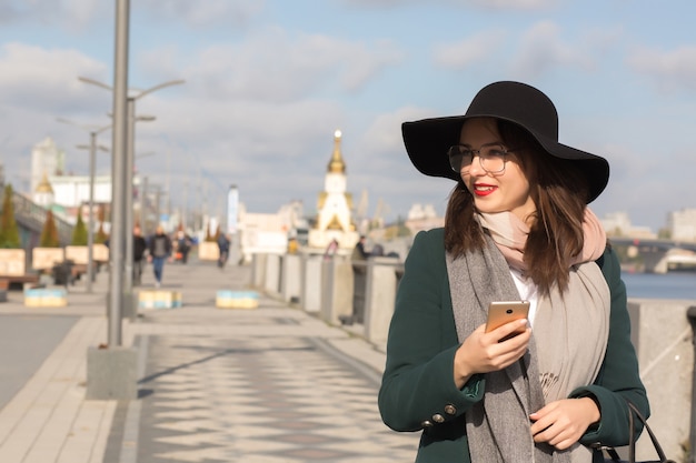 Adorable smiling brunette woman in glasses dressed in stylish apparel typing a message on a mobile phone