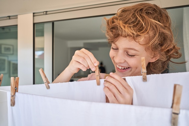 Adorable smiling positive curly haired boy using wooden clothespins for hanging and drying white clean linen after laundry