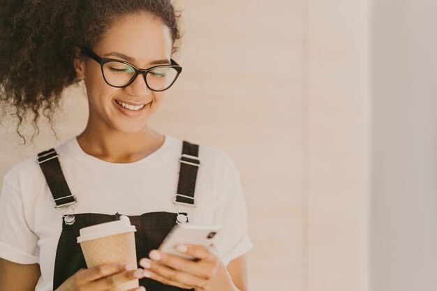 Adorable teeange girl with curly hair checks news feed messages in social networks uses free internet connection wears optical glasses casual white t shirt and overalls drinks takeaway coffee