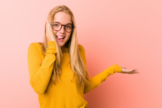 Adorable teenager woman holds copy space on a palm, keep hand over cheek. Amazed and delighted.