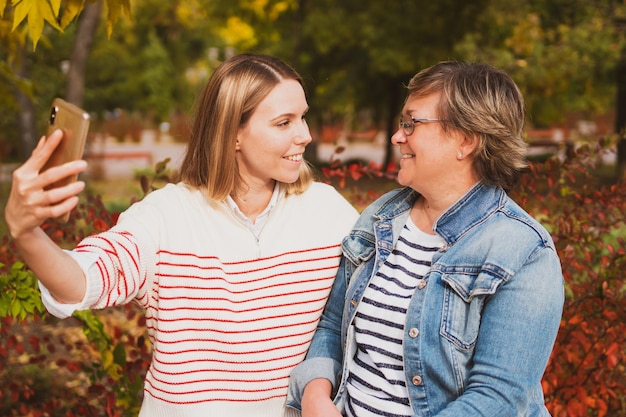 Adorable women mom and daughter are sitting on bench in beautiful autumn park and taking selfie on phone