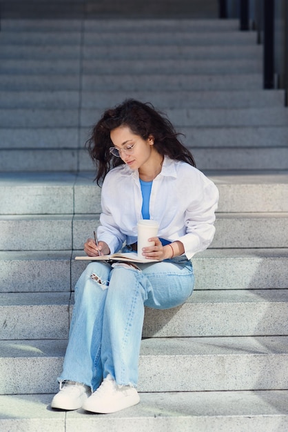 Adorable young office female worker in white shirt with book and coffee sitting on stairs outdoors