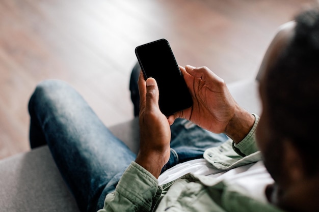 Adult african american man in casual typing on smartphone with blank screen in living room interior