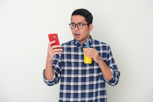 Adult Asian man holding a cup of drink  showing shocked expression when looking to his mobile phone