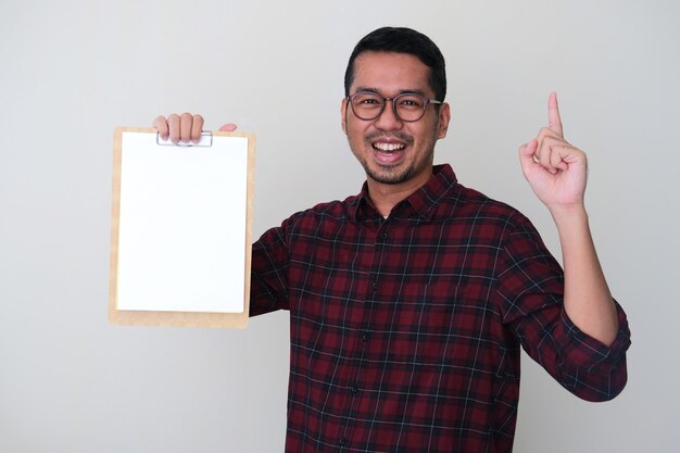 Adult Asian man holding empty white paper in a writing pad with smiling face expression