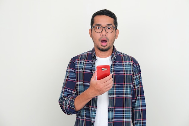 Adult Asian man looking at camera with shocked expression while holding handphone