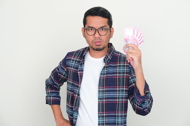 Adult Asian man looking at camera with unhappy expression while holding money