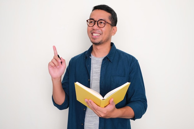 Adult Asian man showing got new idea pose while holding a book and pen