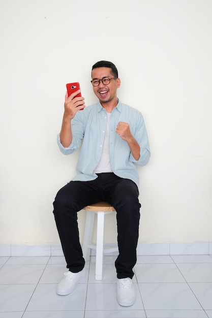 Adult Asian man sitting on a bench looking to his phone with excited expression