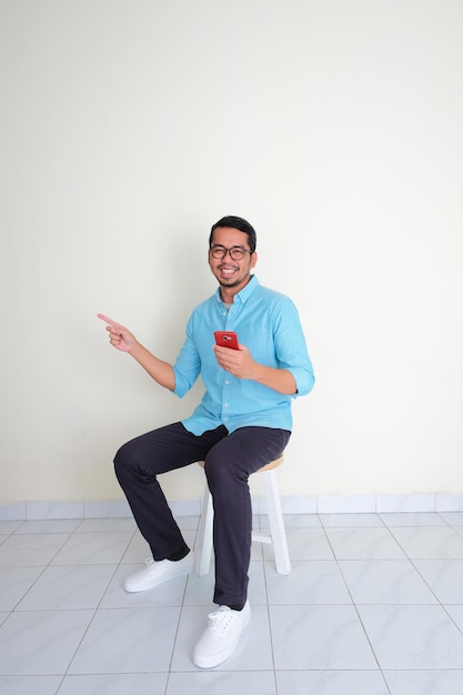 Adult Asian man sitting in a bench pointing to the right side while holding handphone