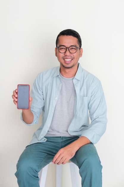 Adult Asian man sitting in a bench showing blank handphone screen with happy expression