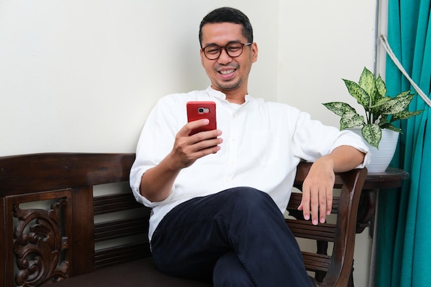 Adult Asian man sitting in a couch smiling when looking to his handphone