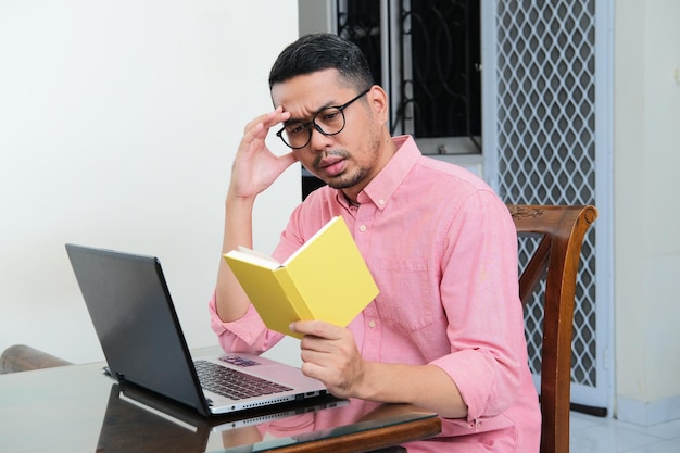 Adult Asian man sitting in front of his laptop while reading a book with serious expression