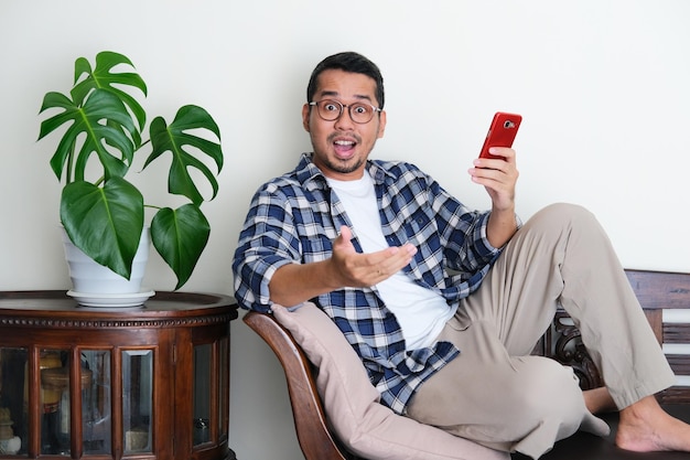 Adult Asian man sitting relax in a couch while holding mobile phone with amazed expression