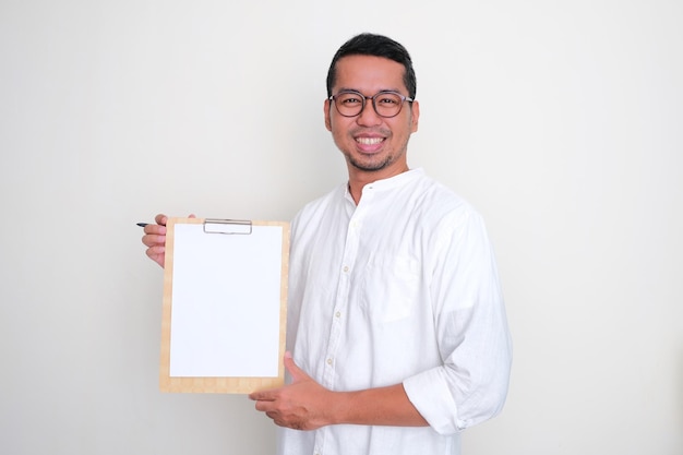 Photo adult asian man smiling confident while showing empty white paper in a clipboard