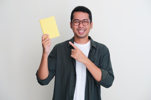 Adult Asian man smiling happy to the camera while pointing to the book that he hold