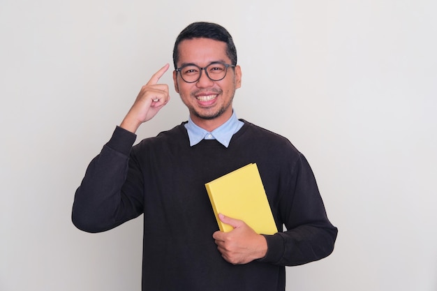 Adult Asian man wearing black sweater smiling while holding a book and pointing to his head