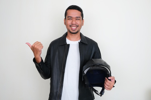 Adult Asian man wearing leather jacket holding motorcycle helmet smiling and pointing to the right side