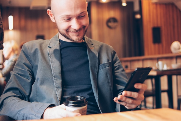 Adult bald smiling man drinking coffee from paper cup and using mobile phone at cafe