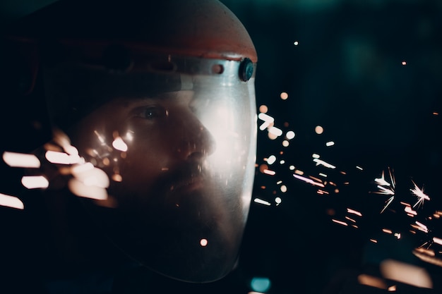 Photo adult bearded man in transparent protective mask and grinder saw with flying metal particles sparks in darkness