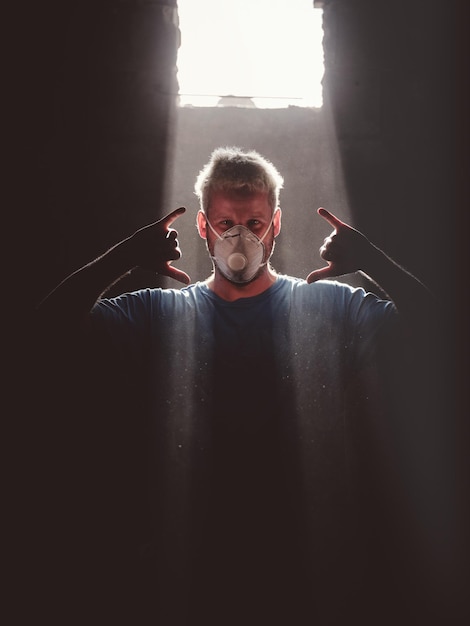 Adult blond haired male surfboard maker in respirator mask showing Hawaiian Shaka gesture while standing near window illuminated with sunbeam in dark room and looking at camera