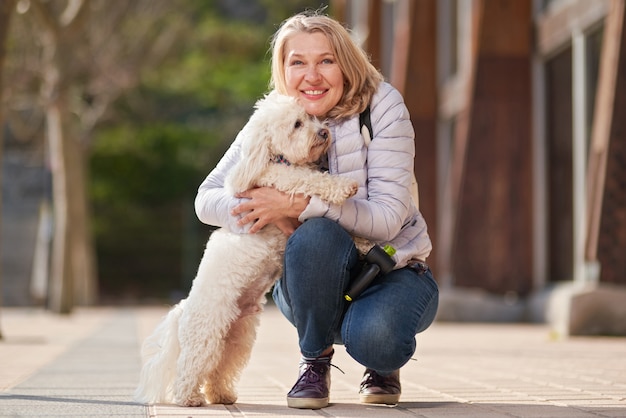 Adult blond woman walking with fluffy white dog in summer city.