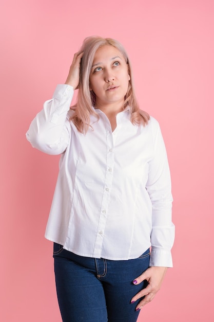 An adult blonde woman scratches her head remembering something while standing on a pink background