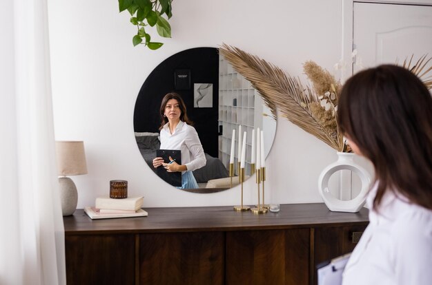 An adult brunette woman in a white blouse is sitting on a chair and looking in the mirror