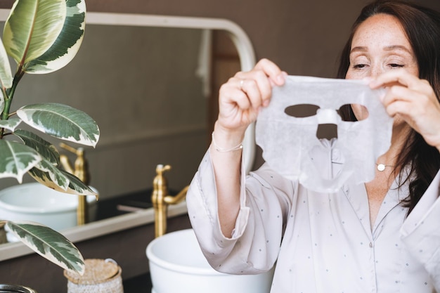 Adult brunette woman with sheet mask on her face in the bathroom at home