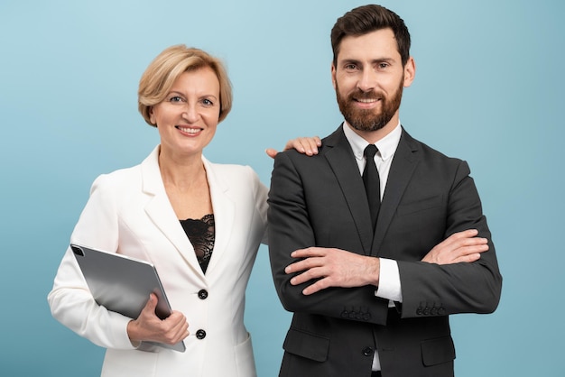 Adult businessman and senior businesswoman wearing formal suits holding laptop posing with pleasure smiles Isolated over blue background