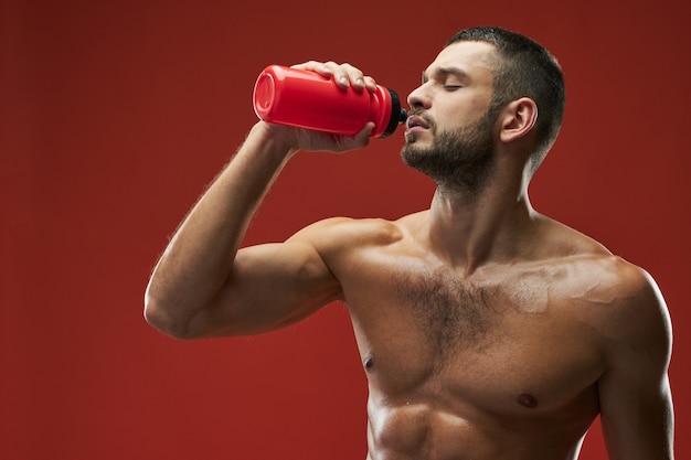 Photo adult caucasian bearded athlete holding red bottle with beverage on red background