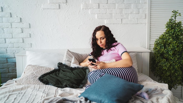 Adult charming female in pajamas flipping through various news in mobile phone in bedroom Young woman learning remotely by smartphone while lying on pillows in bed