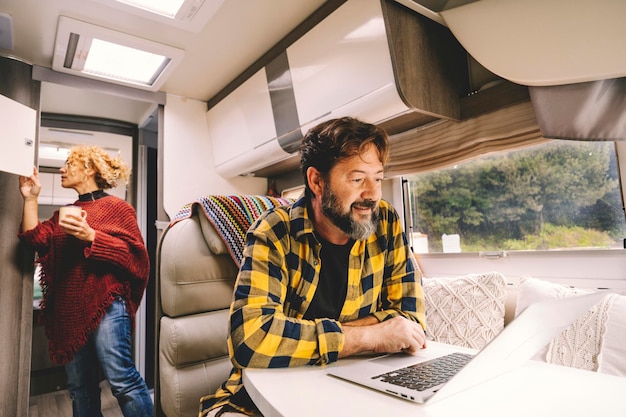 Adult couple travel and enjoy caming car camper van for holiday vacation or vanlife lifestyle - happy man work on laptop on the table and woman looking for stuffs inside the vehicle - nature outdoors