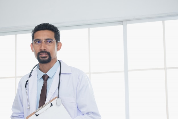 Adult Doctor wearing doctor' gown with a stethoscope hanging on the neck with paperwork standing and looking forward to a reliable manner