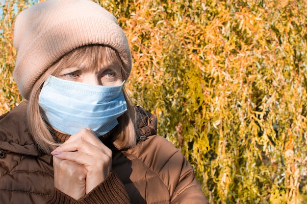 Adult elderly sad senior woman in medical protective mask prays to God in autumn on the street