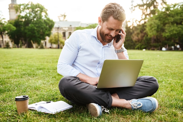 adult european man in business clothing, sitting on grass in park with legs crossed and having business call while working on silver laptop