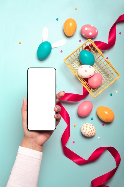 Adult hand holding smartphone with colorful Easter eggs, red ribbon and small yellow basket