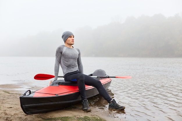 Adult handsome man sitting on boat and looks into distance with dreamily facial expression, wearing sporty wear, posing on foggy river