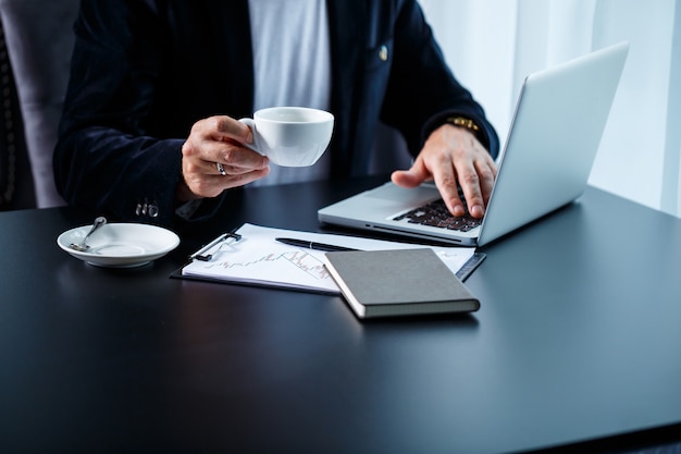 An adult male businessman is working on a new project and looking at the stock growth charts. Sits at the table by the large window. Looks at the laptop screen and drinks coffee.