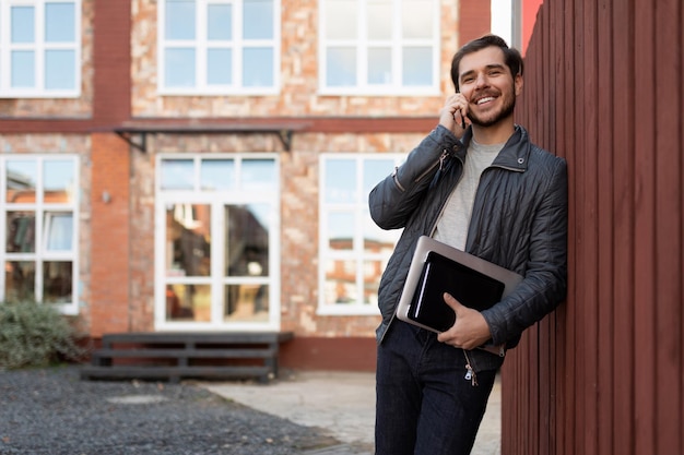 Adult male businessman talking on a mobile phone leaning against a wooden fence next to a brick