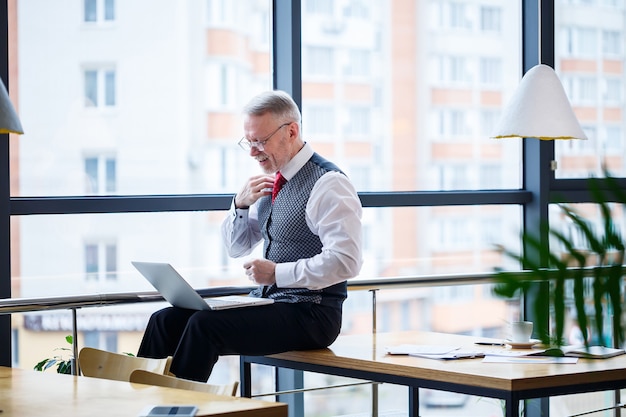 Adult male businessman, teacher, mentor working on a new project. Sits by a large window on the table. He looks at the laptop screen.