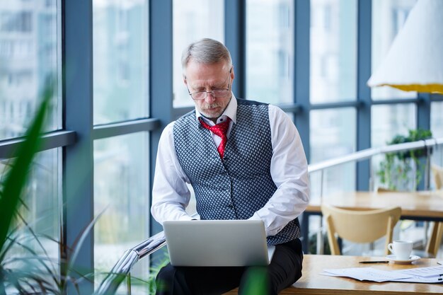 Adult male businessman, teacher, mentor working on a new project. Sits by a large window on the table. He looks at the laptop screen.