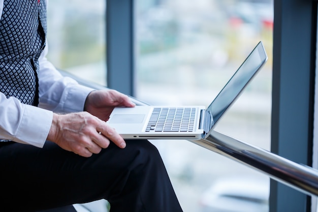 Adult male businessman, teacher, mentor working on a new project. Sits by a large window on the table. He looks at the laptop screen.