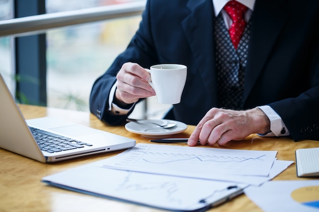 Adult male businessman working on a new project and looking at stock growth charts. Sits at a large window at the table. Looks at the laptop screen and drinks coffee.