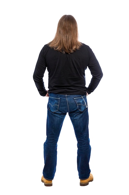 An adult man is standing A guy with long hair in jeans and a black shirt Back view Full height Isolated on white background Vertical