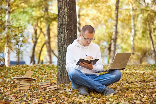 Adult man in a white hoodie is studying in the Park on a laptop, writing in a notebook, reading books and textbooks. Outdoor learning, social distancing